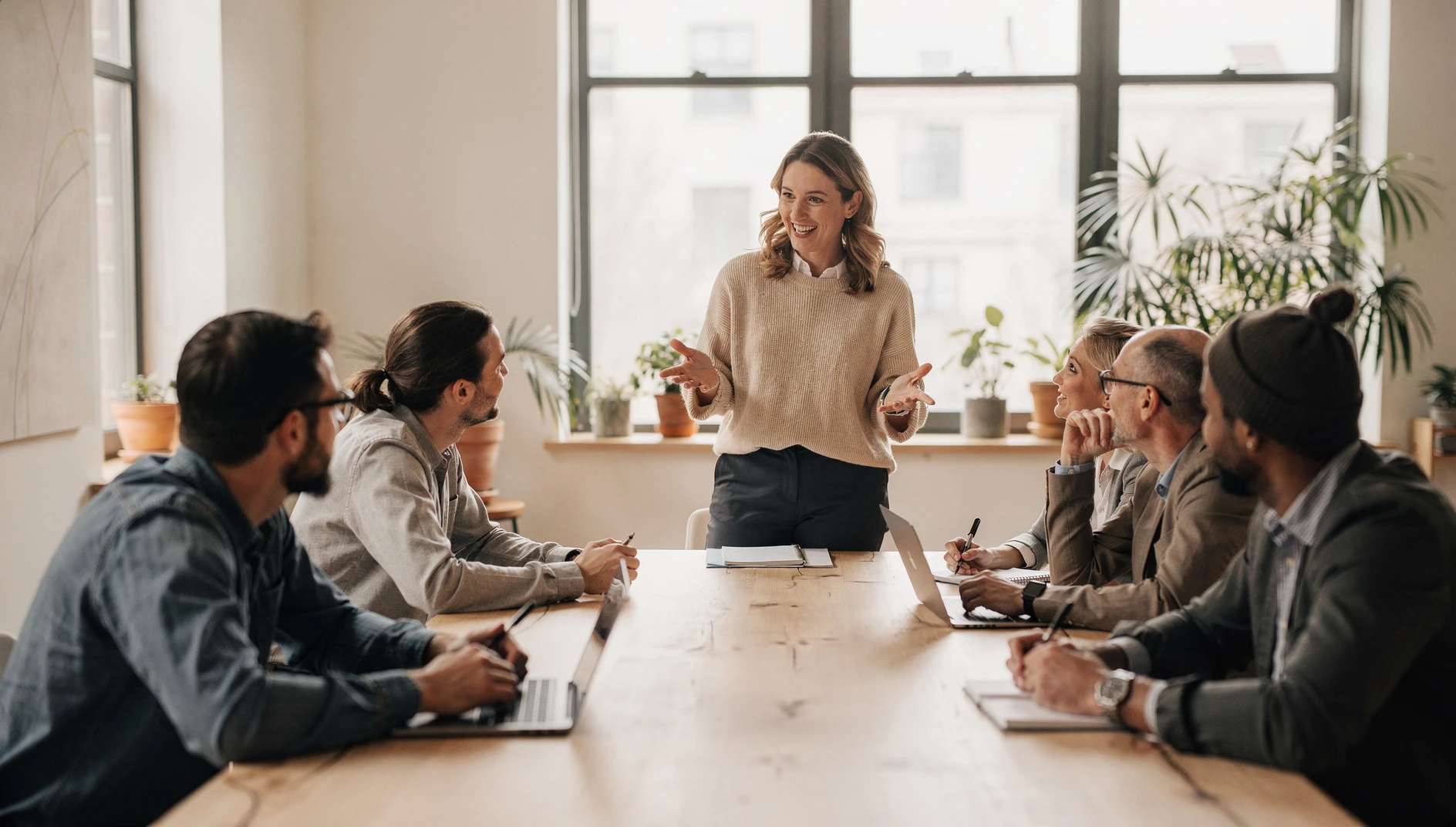 A coach speaking confidently to a small group in a bright workshop room with people seated around a table and engaged expressions in natural light