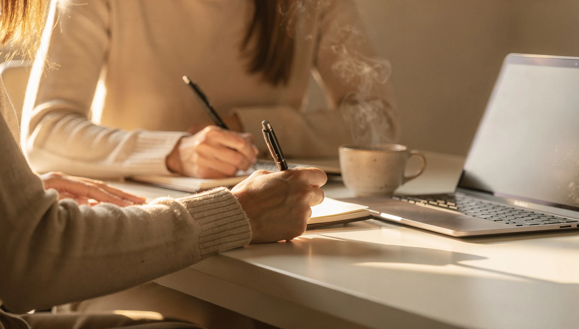 A person writing ideas in a notebook at a bright desk with a laptop open and coffee nearby in afternoon light