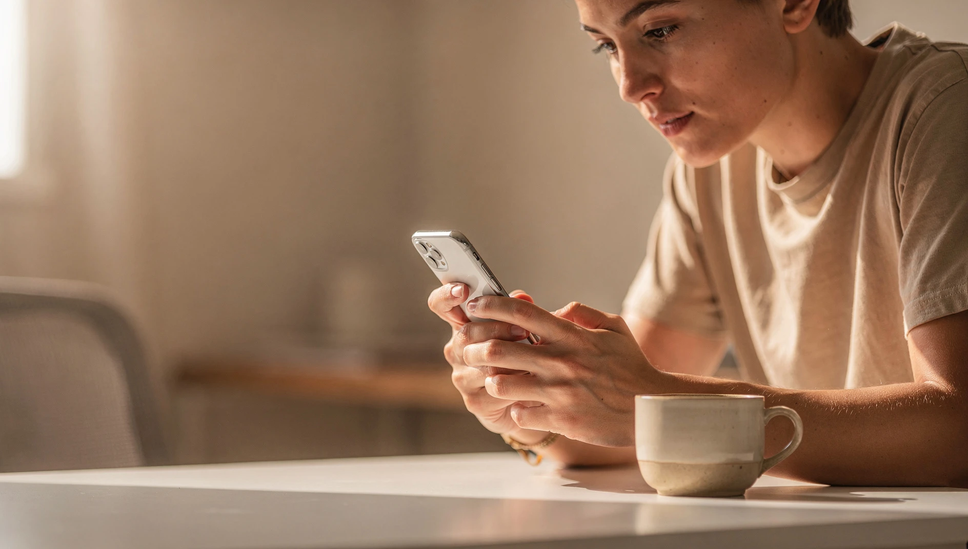 A person looking at a smartphone screen with a focused expression at a bright desk with coffee nearby