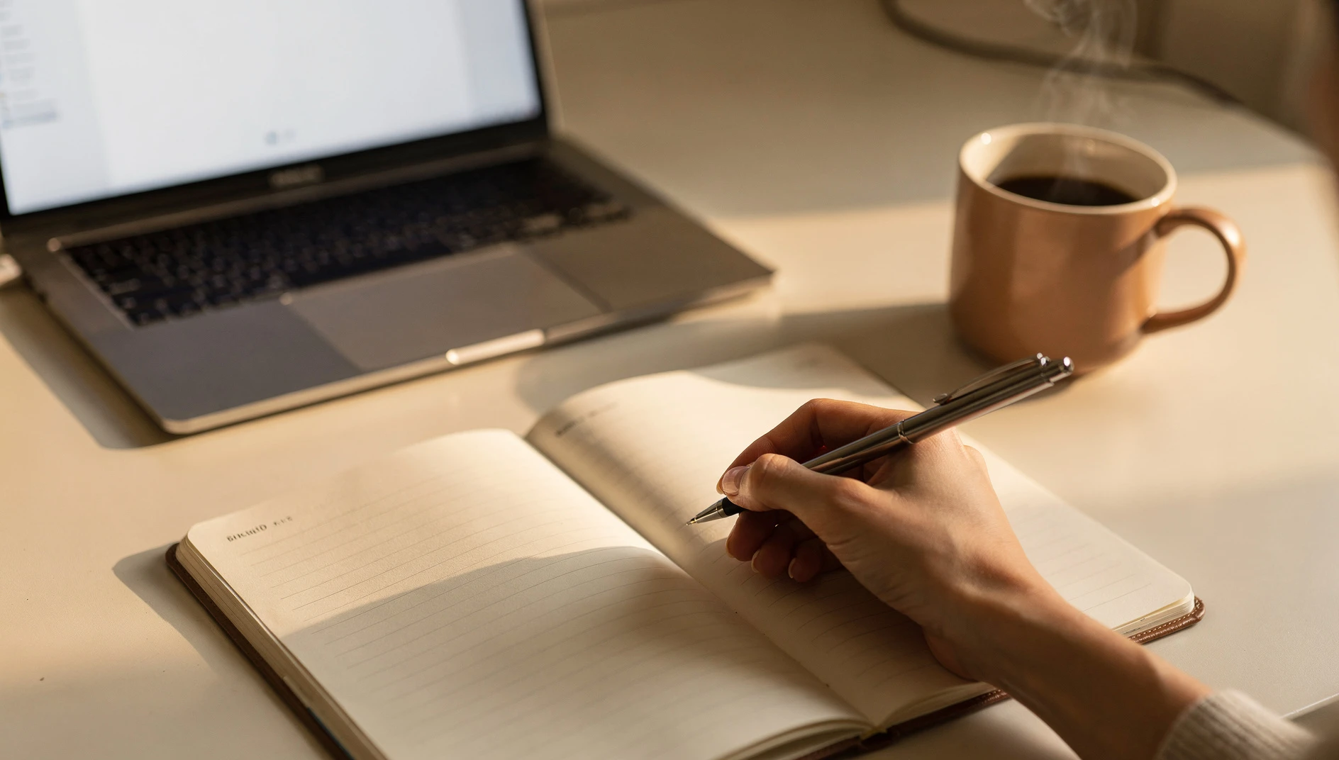 A person writing in a planner at a minimal desk with a laptop and coffee mug in warm morning light